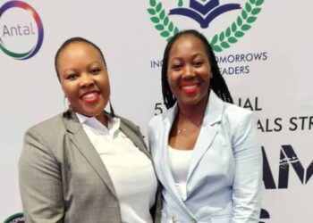 Two professional women in blazer suits smile for the camera at a conference backdrop with sponsor logos, including Antal and others.