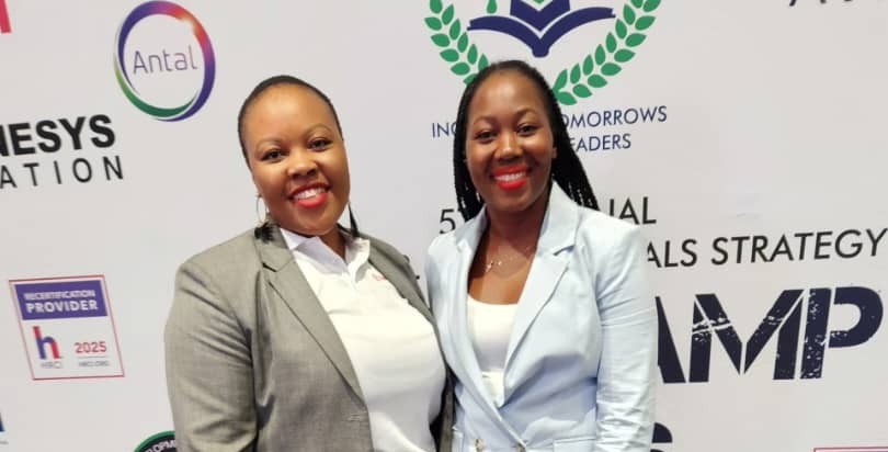 WhatsApp Image 2026-04-16 at 82156 AM - The Brief | Namibia's Leading Business & Financial News Two professional women in blazer suits smile for the camera at a conference backdrop with sponsor logos, including Antal and others.