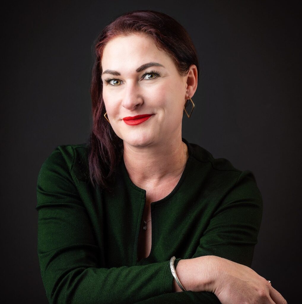Natasja Beyleveld - The Brief | Namibia's Leading Business & Financial News Portrait of a woman with dark red hair, wearing a green blazer, red lipstick, and gold geometric earrings, smiling at the camera against a dark background.