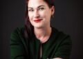 Natasja Beyleveld - The Brief | Namibia's Leading Business & Financial News Portrait of a woman with dark red hair, wearing a green blazer, red lipstick, and gold geometric earrings, smiling at the camera against a dark background.