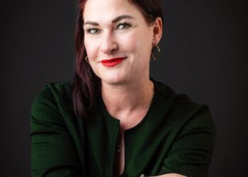 Portrait of a woman with dark red hair, wearing a green blazer, red lipstick, and gold geometric earrings, smiling at the camera against a dark background.
