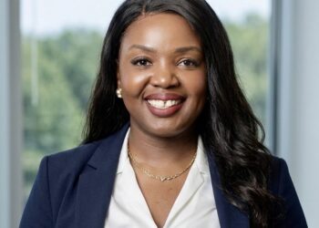 Smiling Black woman wearing a navy blazer and white blouse, in a bright indoor setting with greenery outside the window.