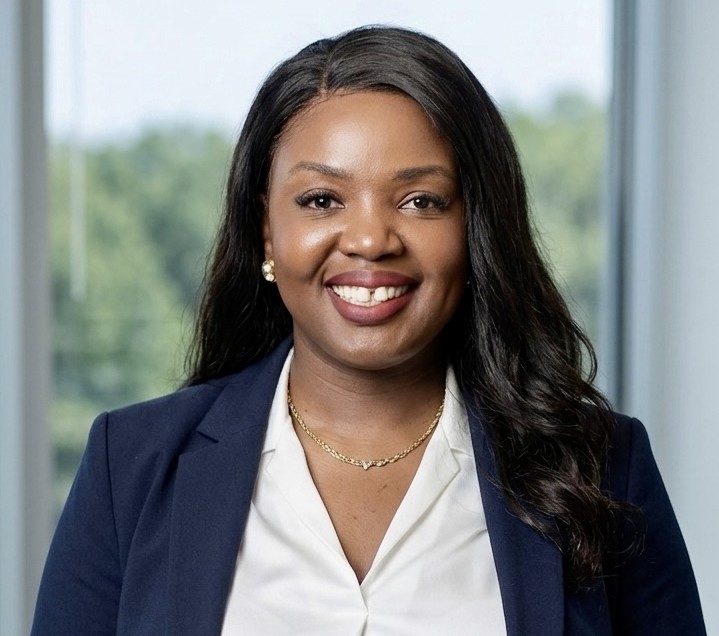 Smiling Black woman wearing a navy blazer and white blouse, in a bright indoor setting with greenery outside the window.