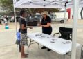 Namibia Deposit Guarantee Fund - The Brief | Namibia's Leading Business & Financial News Two women chat at an outdoor booth under a white canopy, one handing a brochure to a young girl while a table displays rocks and stacks of papers.",