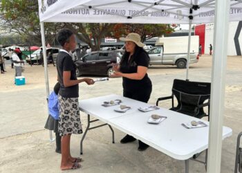 Two women chat at an outdoor booth under a white canopy, one handing a brochure to a young girl while a table displays rocks and stacks of papers.",