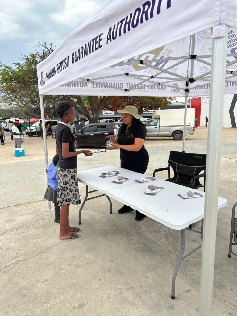 Namibia Deposit Guarantee Fund - The Brief | Namibia's Leading Business & Financial News Two women chat at an outdoor booth under a white canopy, one handing a brochure to a young girl while a table displays rocks and stacks of papers.",