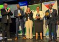 WhatsApp Image 2026-04-16 at 24508 PM - The Brief | Namibia's Leading Business & Financial News Five people on a stage at an awards ceremony, each holding a small golden bowl trophy, with sponsor banners behind.