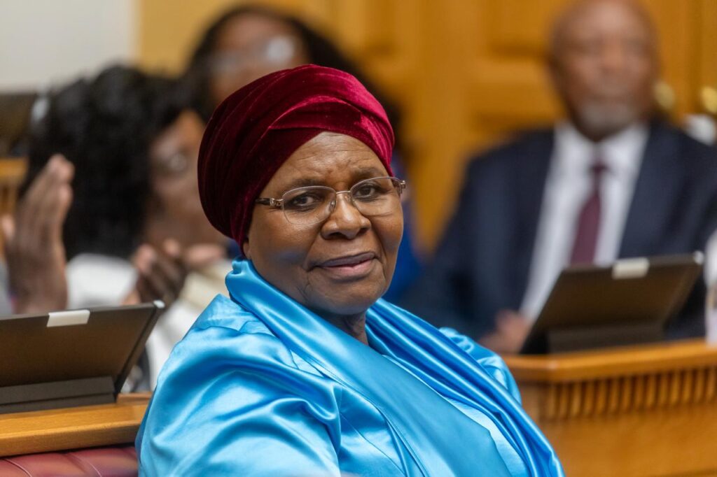 Elderly woman in a blue satin outfit and red headwrap sits in a formal meeting room, smiling at the camera.