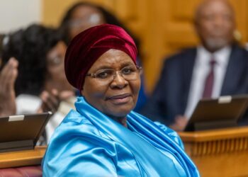 Elderly woman in a blue satin outfit and red headwrap sits in a formal meeting room, smiling at the camera.