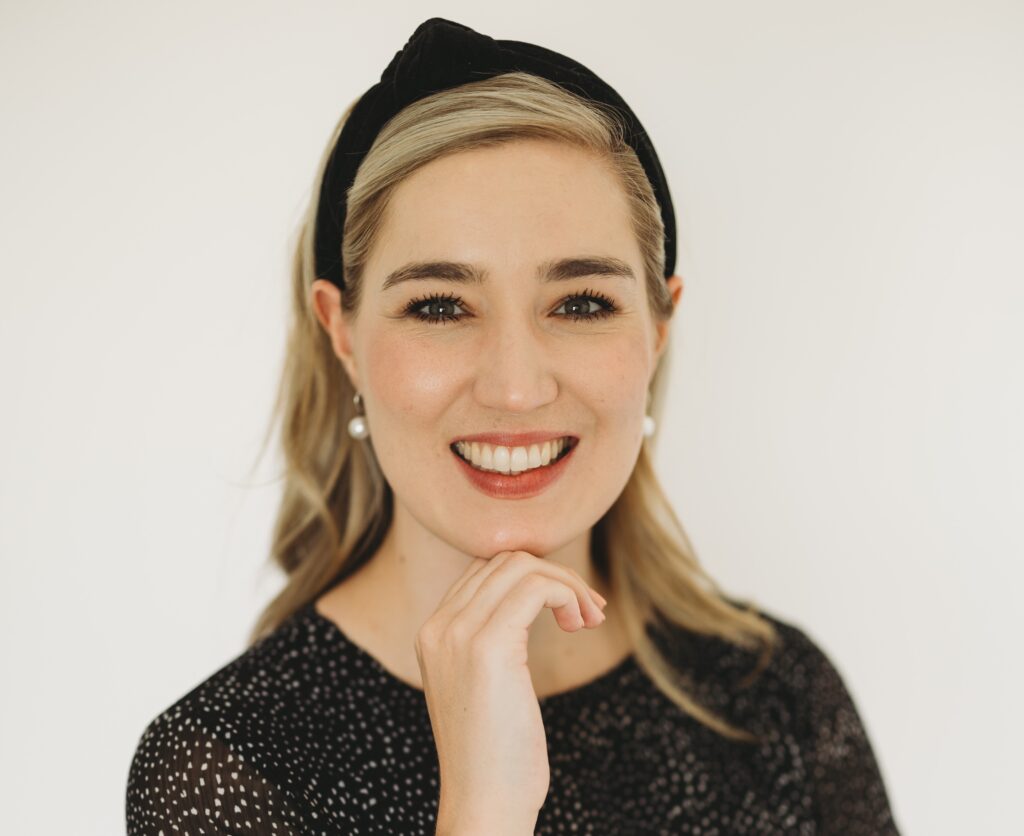 Smiling woman with blonde hair, wearing a black headband and dotted top against a light background, hand resting near chin.