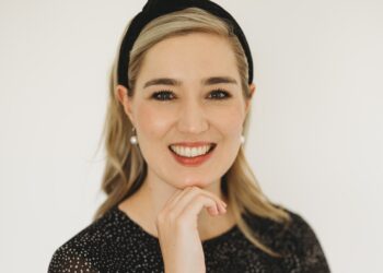 Smiling woman with blonde hair, wearing a black headband and dotted top against a light background, hand resting near chin.