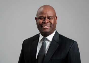 Smiling man in a dark suit and tie against a gray studio backdrop, looking at the camera, professional headshot.