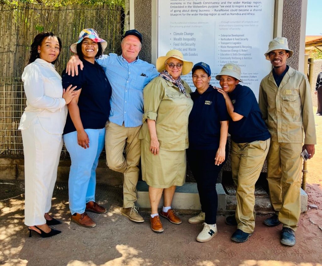 Group of seven smiling people posing arm-in-arm outdoors in front of an information sign; mix of uniforms and casual clothing indicating volunteers or staff.