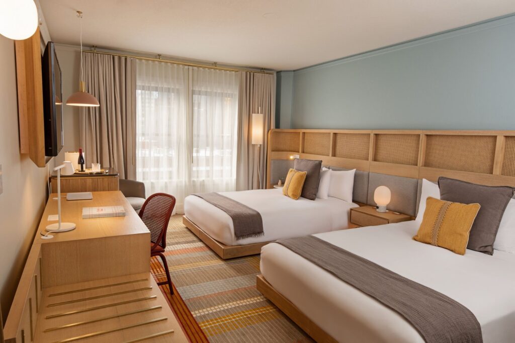 WhatsApp Image 2026-04-17 at 42200 PM - The Brief | Namibia's Leading Business & Financial News Hotel room with two twin beds, a light wood headboard, and pastel blue wall; desk, chair, and a window with sheer curtains in the background.
