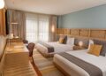Hotel room with two twin beds, a light wood headboard, and pastel blue wall; desk, chair, and a window with sheer curtains in the background.