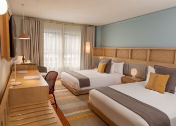 Hotel room with two twin beds, a light wood headboard, and pastel blue wall; desk, chair, and a window with sheer curtains in the background.