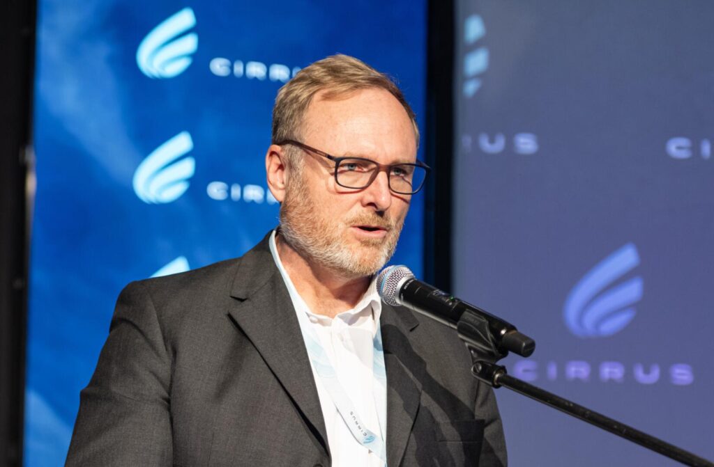 Middle-aged man with glasses in a gray suit speaks into a microphone at a conference, with a blue branded backdrop behind him.