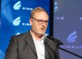 JOHN ANDRES - The Brief | Namibia's Leading Business & Financial News Middle-aged man with glasses in a gray suit speaks into a microphone at a conference, with a blue branded backdrop behind him.
