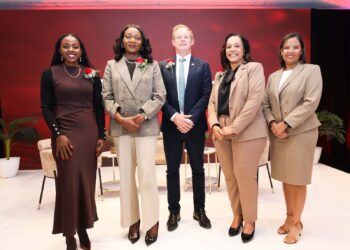 Five professionals pose on a stage with a red backdrop, wearing business attire and corsages on their lapels.