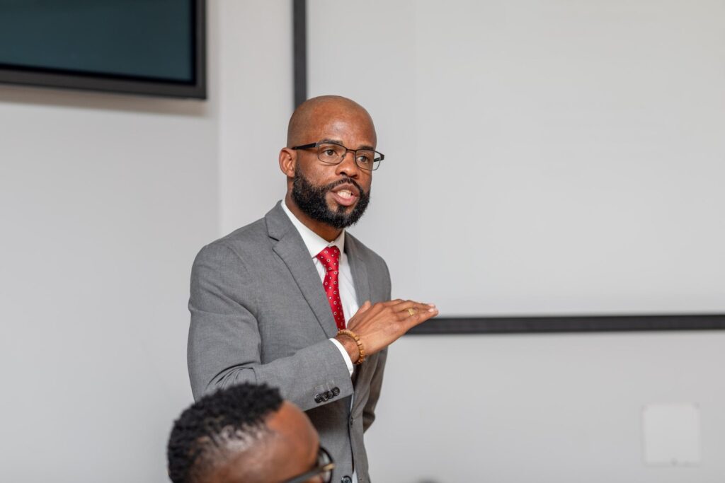 STANLEY SHANAPINDA2 - The Brief | Namibia's Leading Business & Financial News Man in a gray suit and red tie speaks, gesturing with his hands during a presentation in a meeting room.