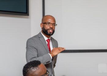 Man in a gray suit and red tie speaks, gesturing with his hands during a presentation in a meeting room.