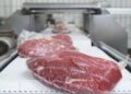 Raw red meat wrapped in plastic on a conveyor belt in a meat processing facility, foreground in focus.