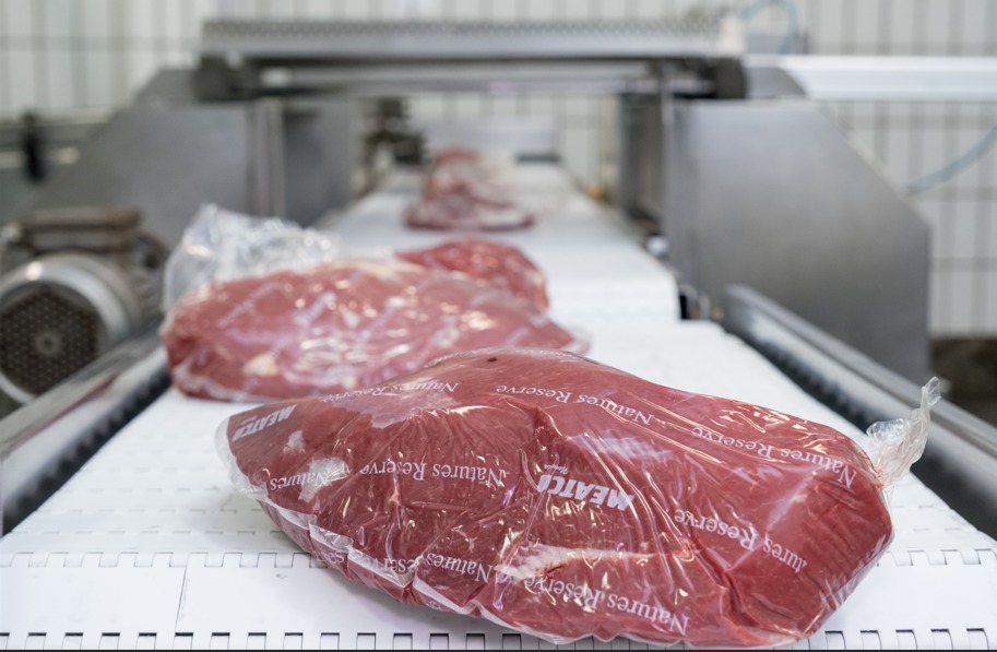 Raw red meat wrapped in plastic on a conveyor belt in a meat processing facility, foreground in focus.