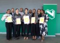 Group of eight professionals posing with framed certificates at a ceremony, in front of a green wall with a logo behind them.