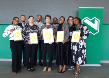 Group of eight professionals posing with framed certificates at a ceremony, in front of a green wall with a logo behind them.