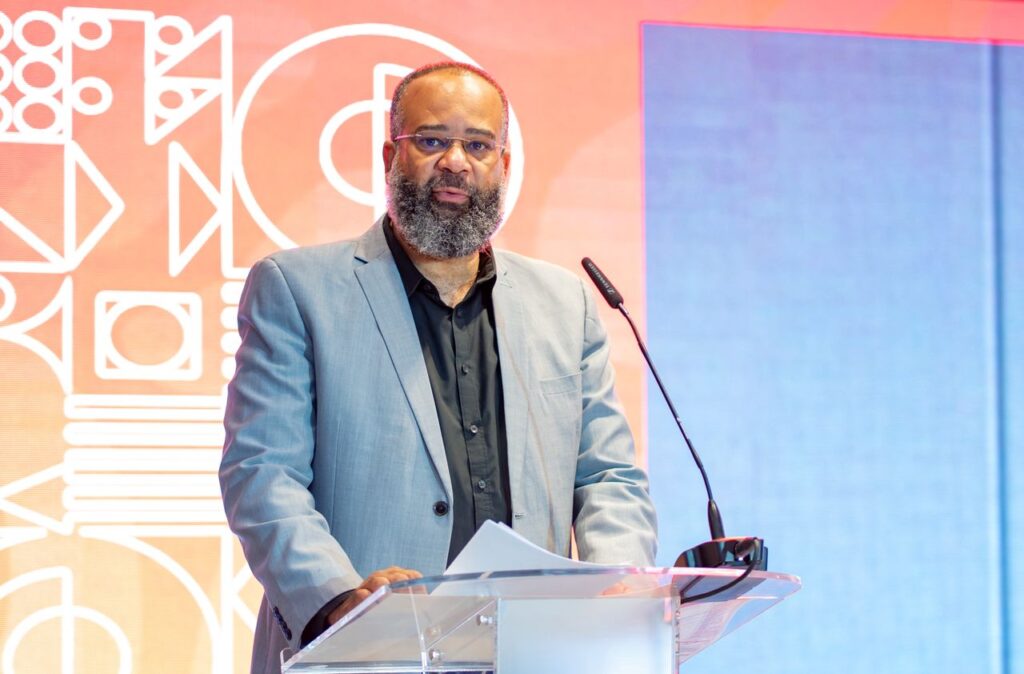 CARLO - The Brief | Namibia's Leading Business & Financial News Man in a gray suit delivers a speech at a clear podium with a microphone, against an orange and blue geometric backdrop.
