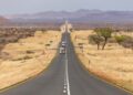ROAD - The Brief | Namibia's Leading Business & Financial News Long straight desert highway stretching to distant mountains with a few cars traveling in single file, signposts along the road.