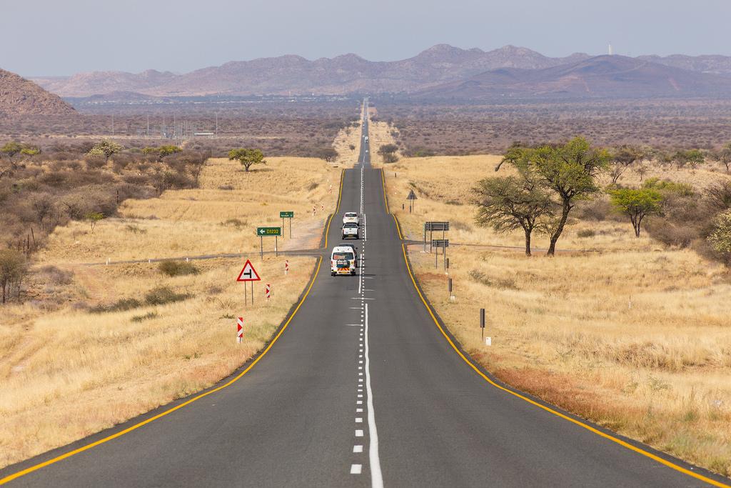 ROAD - The Brief | Namibia's Leading Business & Financial News Long straight desert highway stretching to distant mountains with a few cars traveling in single file, signposts along the road.