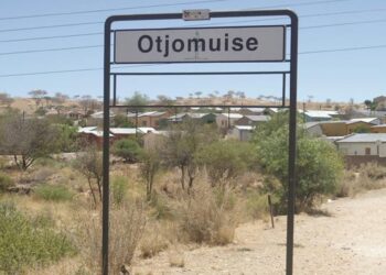 Town entrance sign reading 'Otjomuse' over a dusty, dry landscape with small houses in the background.