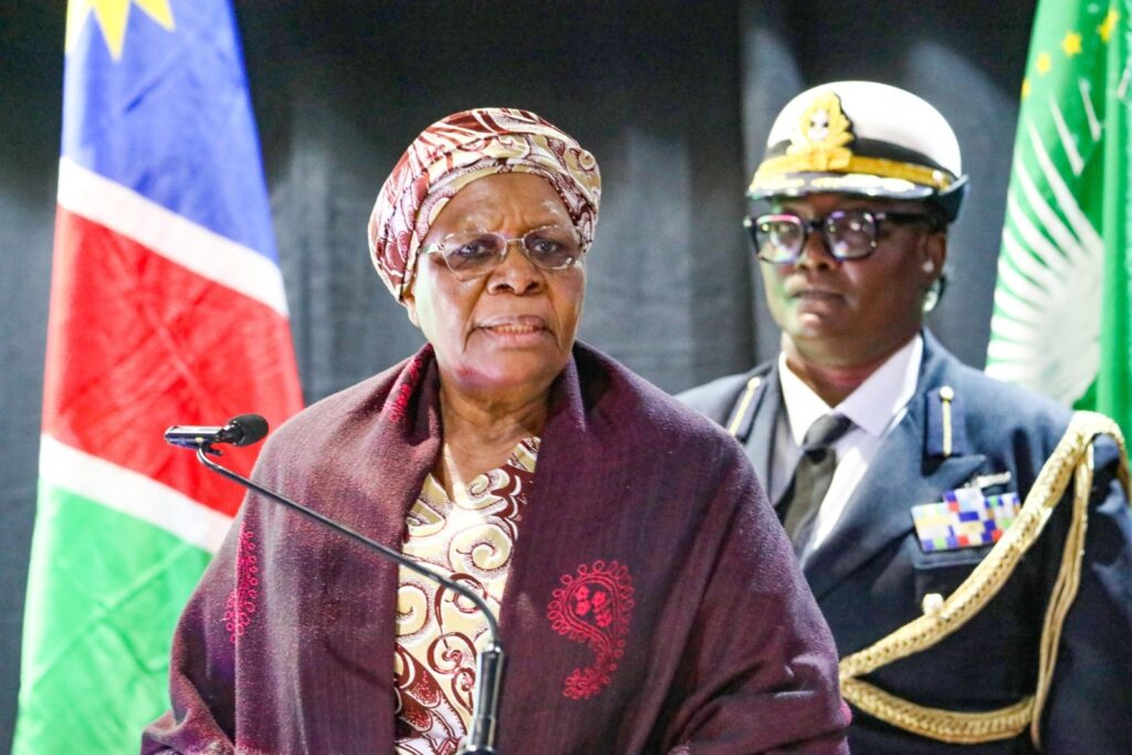 Senior woman in a patterned headwrap and glasses speaks at a podium with a microphone, a flag behind her and a uniformed officer in the background.