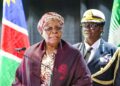 WhatsApp Image 2026-04-22 at 54740 PM - The Brief | Namibia's Leading Business & Financial News Senior woman in a patterned headwrap and glasses speaks at a podium with a microphone, a flag behind her and a uniformed officer in the background.