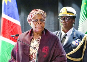 Senior woman in a patterned headwrap and glasses speaks at a podium with a microphone, a flag behind her and a uniformed officer in the background.