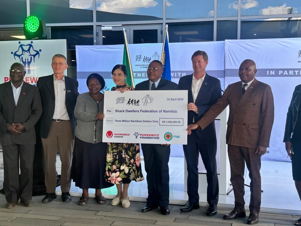 Group of professionals in formal attire posing with a large donation check during a ceremony, banners and flags in the background.