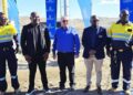 Group of five men posing for a photo at a construction or mining site, two in high-visibility uniforms, flags in the background