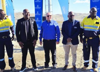 Group of five men posing for a photo at a construction or mining site, two in high-visibility uniforms, flags in the background