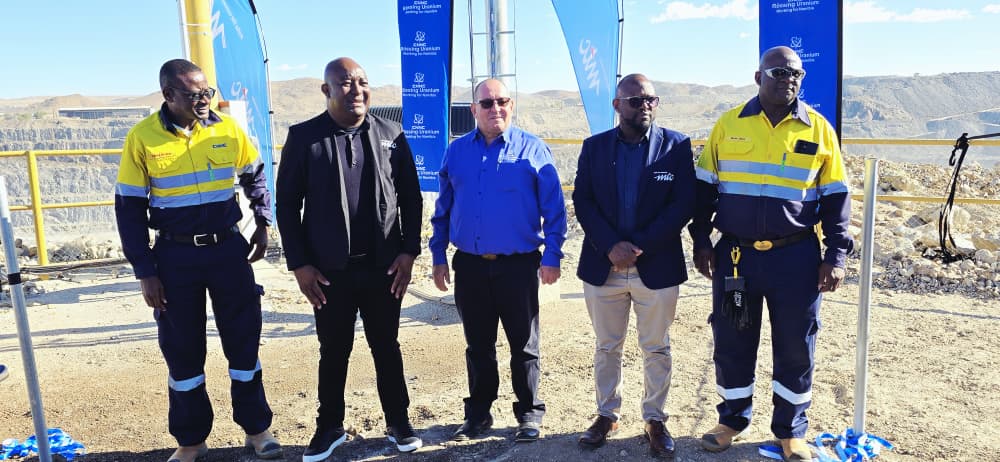 WhatsApp Image 2026-04-23 at 40555 PM - The Brief | Namibia's Leading Business & Financial News Group of five men posing for a photo at a construction or mining site, two in high-visibility uniforms, flags in the background