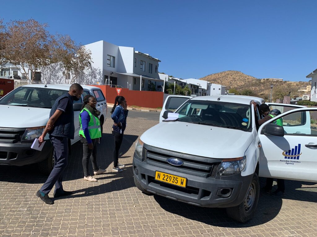 People stand by white vans with Namibia Statistics Agency logo in a sunny residential area. A man in a dark vest, a woman in a green safety vest, and another woman converse nearby.