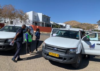 People stand by white vans with Namibia Statistics Agency logo in a sunny residential area. A man in a dark vest, a woman in a green safety vest, and another woman converse nearby.
