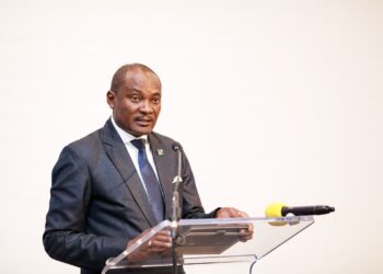 Man in a dark suit and blue tie delivering a speech at a clear lectern with microphones.