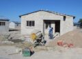 Construction site with a partially built concrete block house, a cement mixer, and a worker moving sand under a clear blue sky.]?