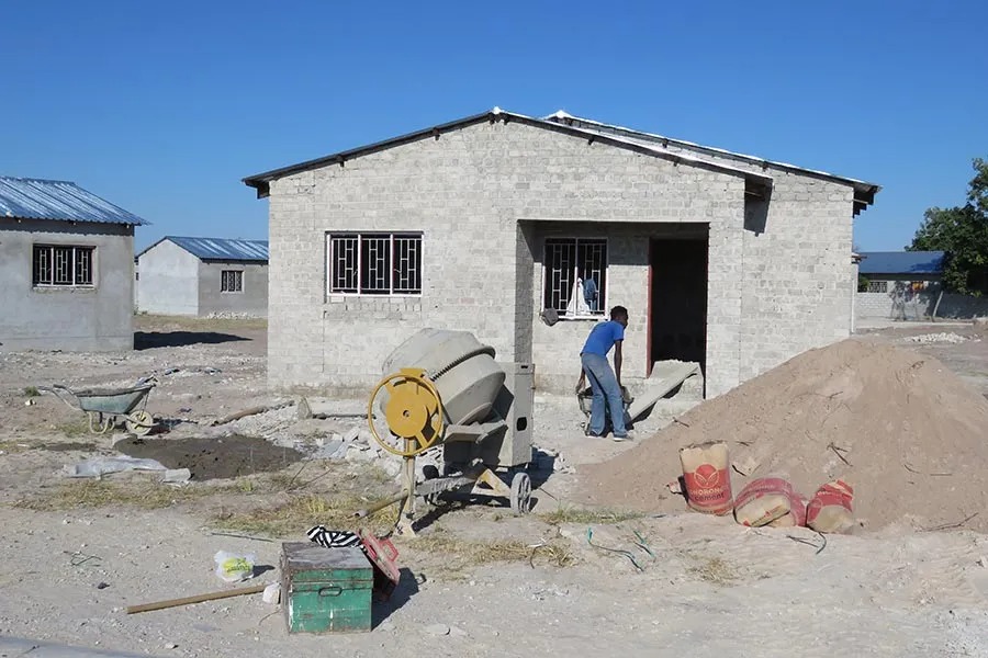 Construction site with a partially built concrete block house, a cement mixer, and a worker moving sand under a clear blue sky.]?