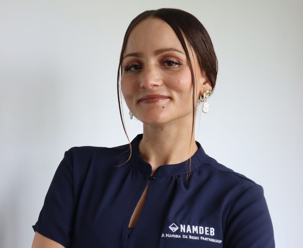 Portrait of a woman in a navy blouse, facing the camera, wearing a NAMDEB logo shirt with a beaded earring visible on the right ear, neutral background.