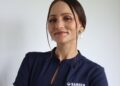 Portrait of a woman in a navy blouse, facing the camera, wearing a NAMDEB logo shirt with a beaded earring visible on the right ear, neutral background.