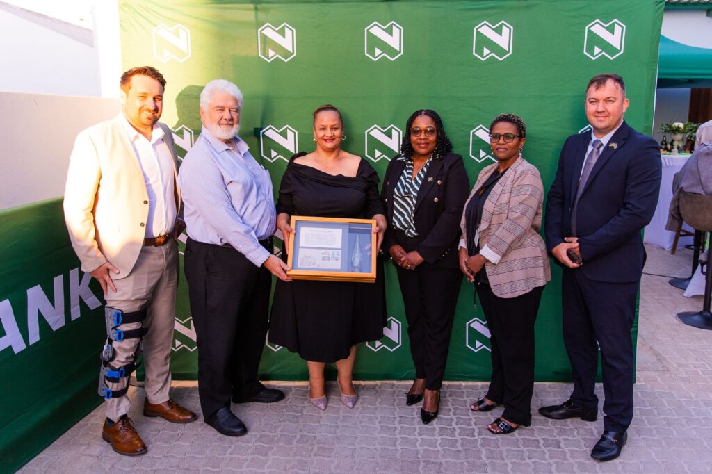 WhatsApp Image 2026-04-27 at 103902 AM - The Brief | Namibia's Leading Business & Financial News Group of six adults posing for a photo at an award ceremony; a woman in black holds a framed certificate with two people beside her. A green branded backdrop is behind them.
