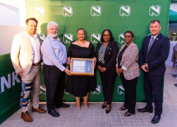 Group of six adults posing for a photo at an award ceremony; a woman in black holds a framed certificate with two people beside her. A green branded backdrop is behind them.