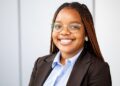 Professional headshot of a smiling Black woman with braided hair, wearing glasses, a light blue shirt and black blazer in an office setting.